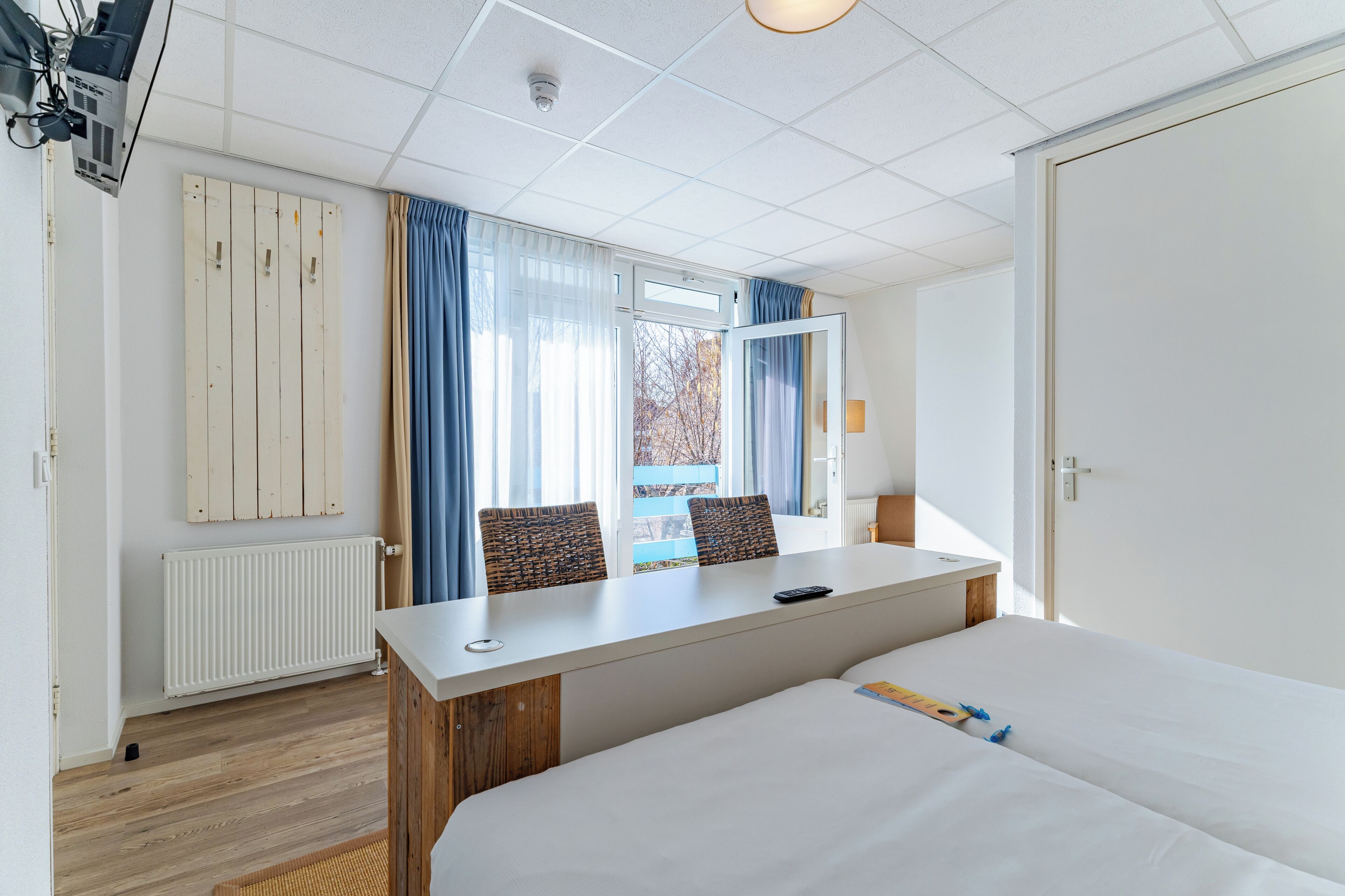 A hotel room with a bed, a desk with two chairs, a radiator, and a window with curtains.
