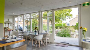 A modern dining area with a glass wall, wooden flooring, and a view of the garden and buildings outside. - Hotel Kijkduin (Domburg)