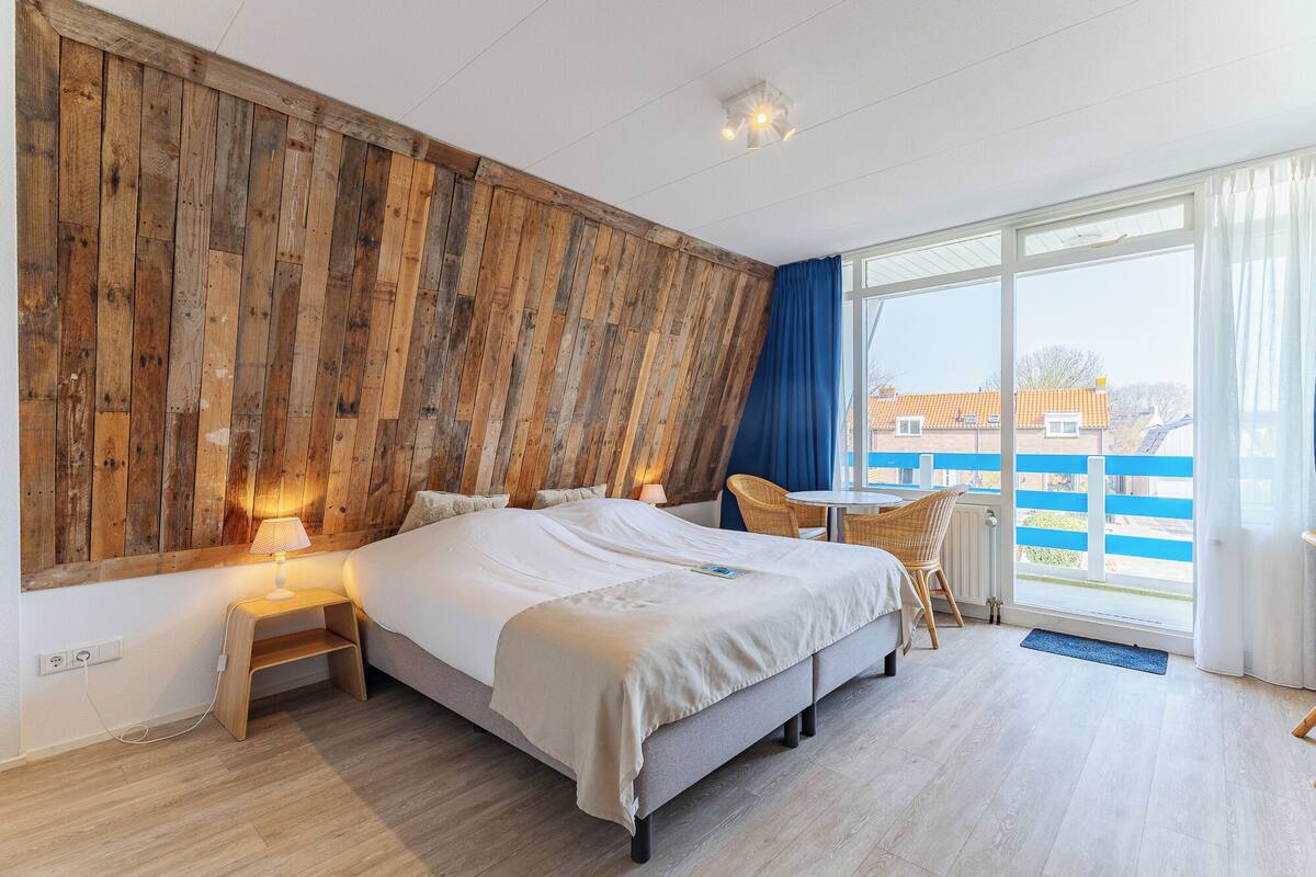A bedroom with a wooden wall, a bed, a chair, a table, and a view of the outside.