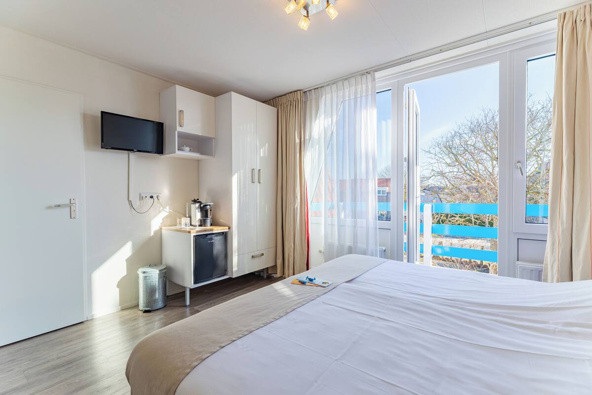 A modern hotel room with a bed, a television, a kitchenette, and a sliding glass door to the outside.
