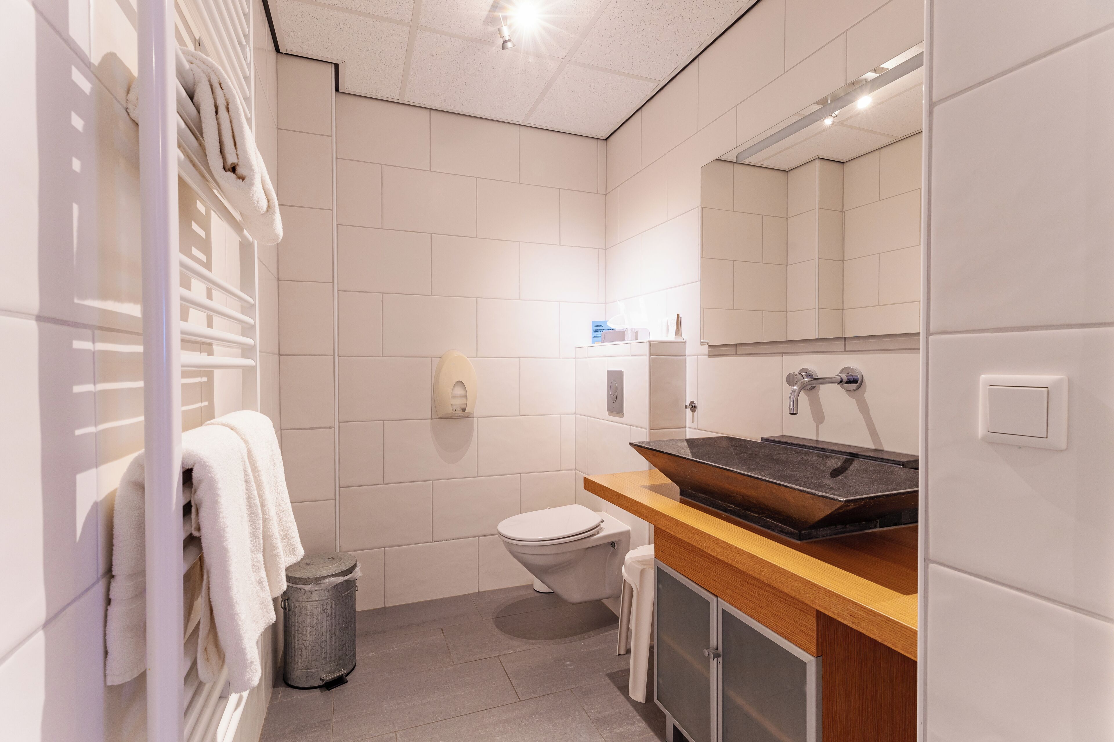 A modern bathroom with a white tiled wall, a wooden vanity with a sink, a toilet, and a towel rack with towels.