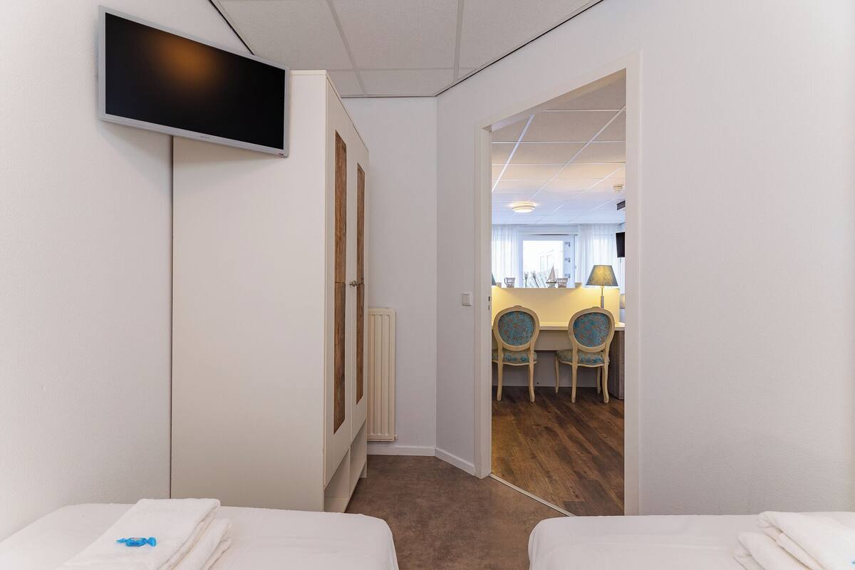 A hotel room with two beds, a TV mounted on the wall, and a view into the dining area.
