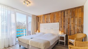 A bedroom with a bed, bedside tables, a chair, a wooden wall, and a window. - Hotel Kijkduin (Domburg)