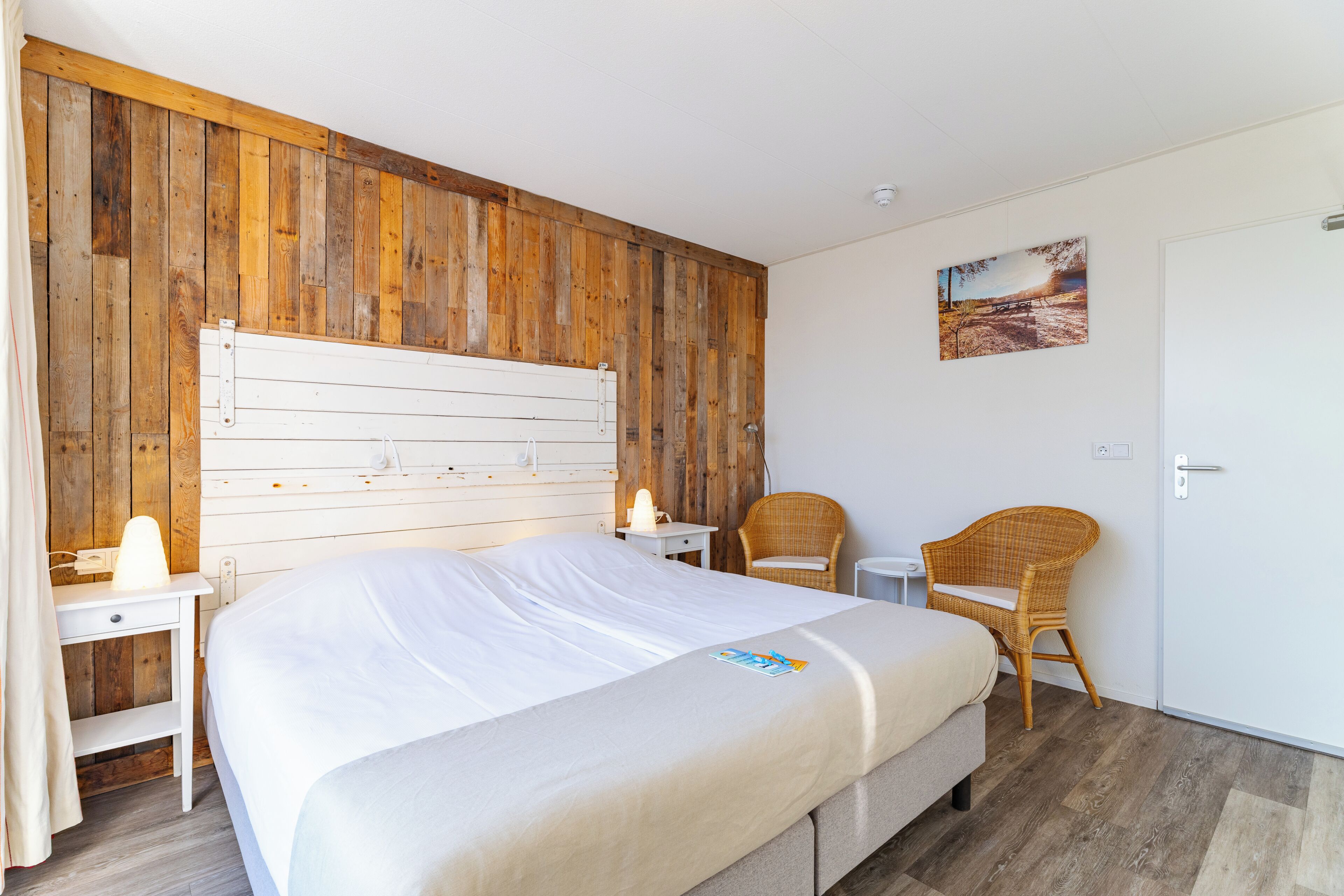 A bedroom with a wooden headboard, a bed, two chairs, a small table, and a painting on the wall.
