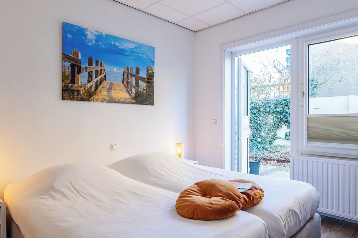 A bedroom with two beds, a painting of a beach scene, and a window with a view of greenery.