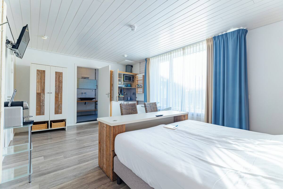 A modern hotel room with a bed, a desk, a wardrobe, and a kitchenette.