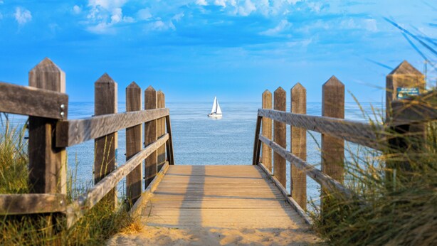 A wooden boardwalk leading to a beach with a sailboat in the distance. - Hotel Kijkduin (Domburg)