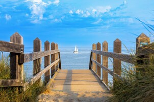 Ein hölzerner Steg führt zu einem Strand, im Hintergrund ein Segelboot.