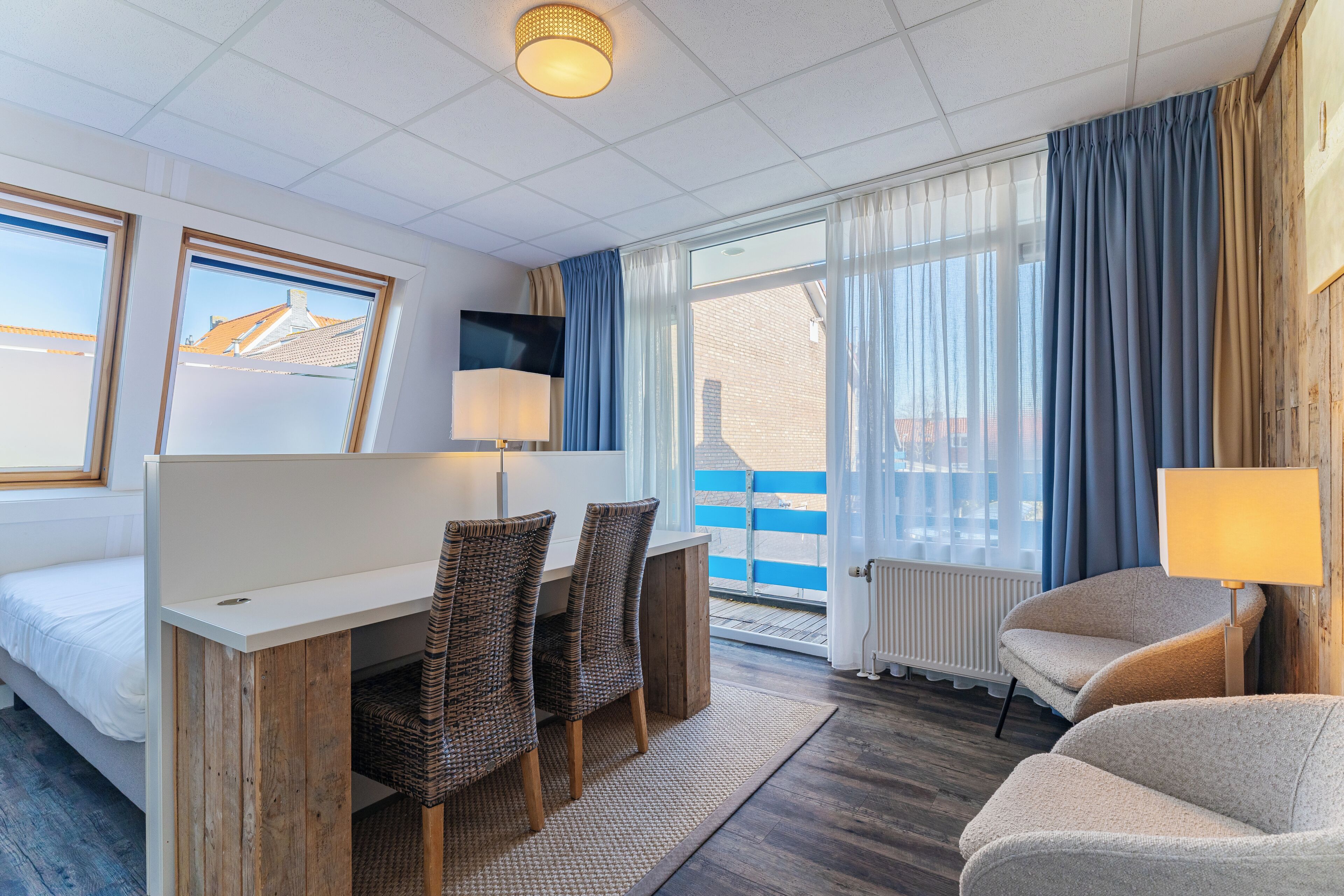 Habitación de hotel con cama, escritorio con sillas, televisor, lámpara y ventanas amplias con cortinas.