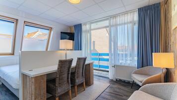 Habitación de hotel con cama, escritorio con sillas, televisor, lámpara y ventanas amplias con cortinas.