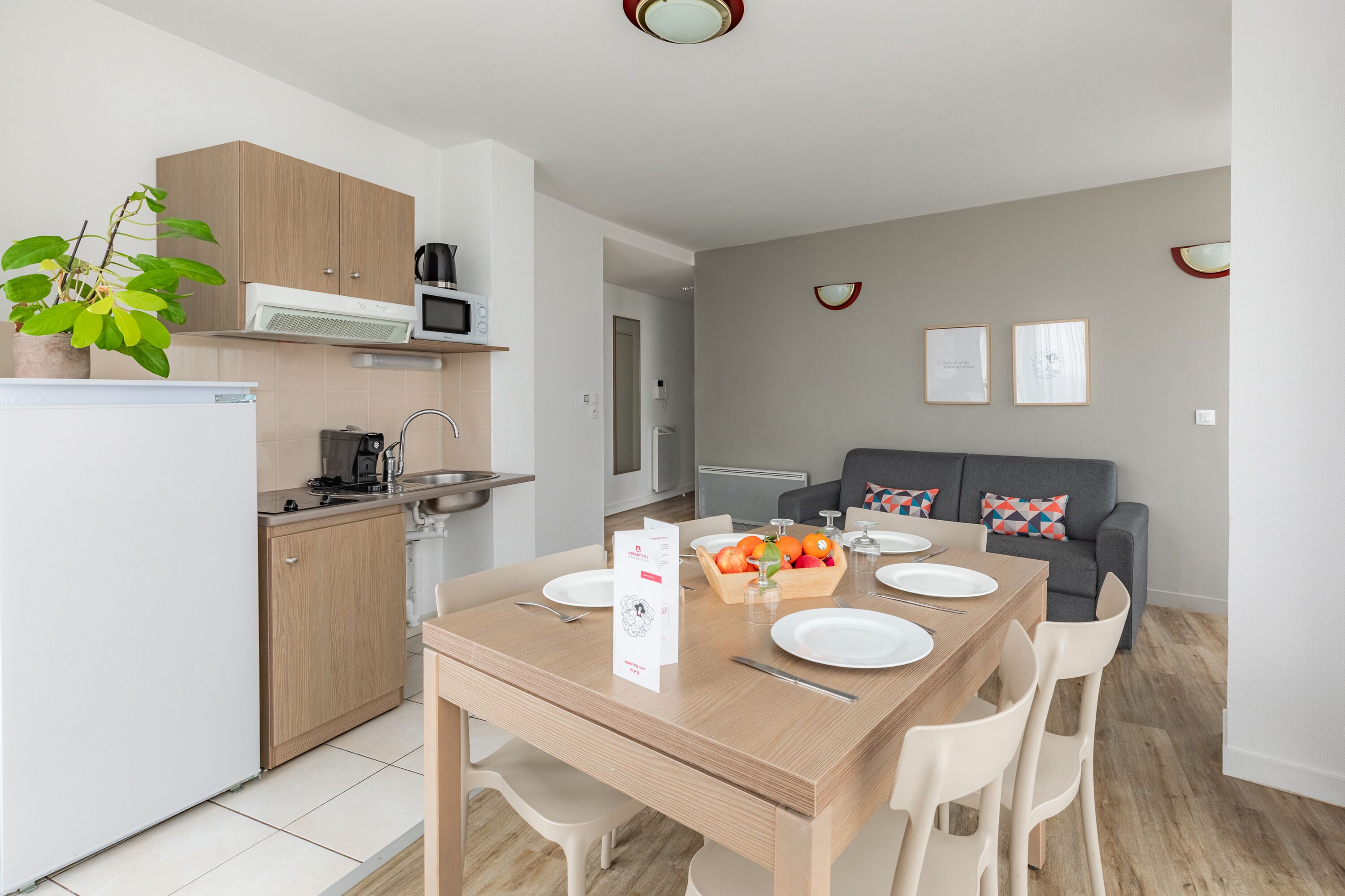 Una cocina moderna con microondas, estufa y armarios. Hay una mesa de comedor con sillas y una cesta de frutas. Un sofá gris con almohadones de colores se ve al fondo.