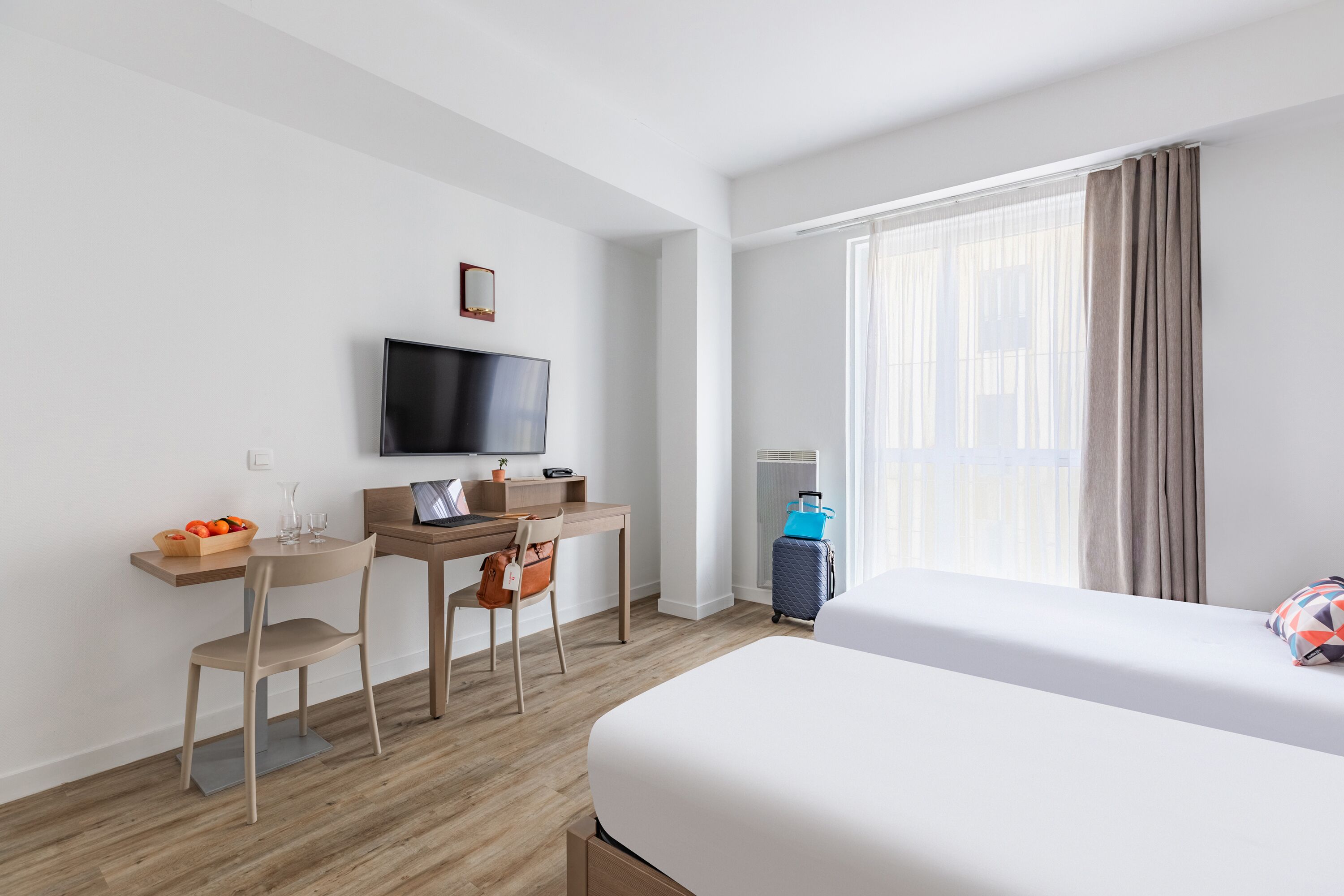 Une chambre d’hôtel comprenant un lit, un bureau avec une chaise, une télévision et une fenêtre avec des rideaux.