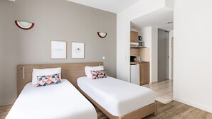 Habitación de hotel con dos camas individuales, cabecero de madera, una pequeña cocina y una puerta que da al baño.