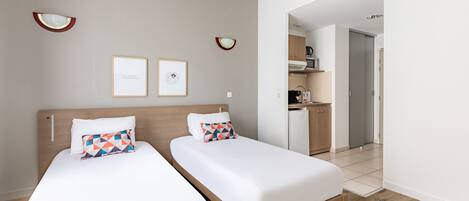 A hotel room with two single beds, a wooden headboard, a small kitchenette, and a door leading to a bathroom.