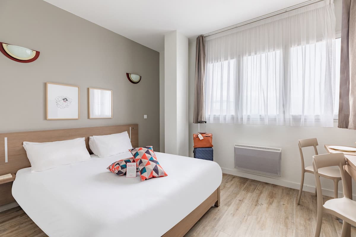Ein modernes Hotelzimmer mit einem großen Bett, einem Schreibtisch und einem Stuhl. An der Wand hängen zwei gerahmte Bilder, ein Fenster mit leichtem Vorhang und ein Heizkörper darunter.