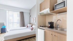 Habitación de hotel con cama, kitchenette, microondas, cafetera y maleta.