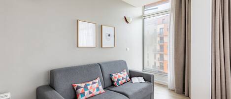 Una sala de estar moderna con un sofá gris, dos cojines geométricos de colores y un ventanal con cortinas.