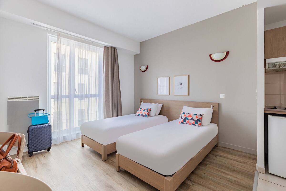 Ein Hotelzimmer mit zwei Betten, einem Schreibtisch, einem Stuhl, einem Koffer und einer Küchenzeile.
