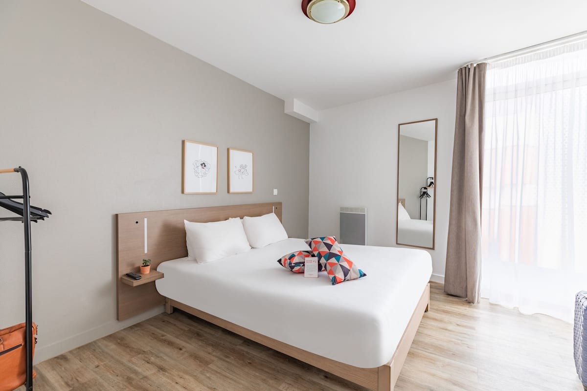 Ein Hotelzimmer mit einem großen Bett, einem Nachttisch, einem an der Wand befestigten Regal, einem Spiegel und einem Fenster mit Vorhängen.