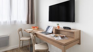 Habitación de hotel moderna con un escritorio de madera, dos sillas, una laptop, un televisor de pantalla plana en la pared, una planta pequeña en maceta y un frutero sobre el escritorio.