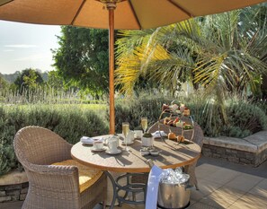 A round table with a wicker chair, a wicker basket, a metal pitcher, and a glass of champagne.