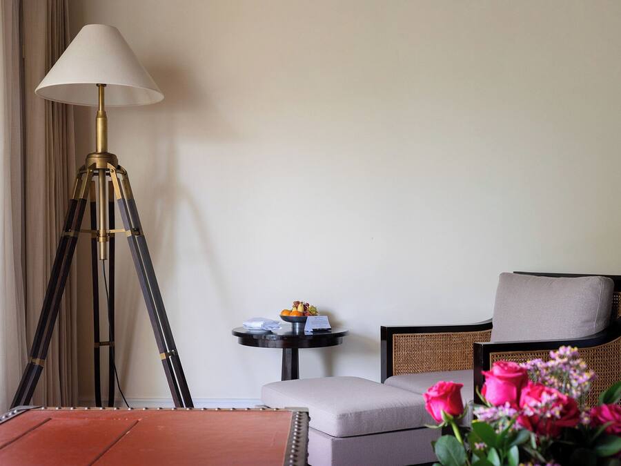 Una habitación con una lámpara de pie, una mesa redonda con un frutero pequeño, un sillón acolchado y un jarrón de flores.