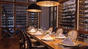 A dining area with a long wooden table set for a meal, surrounded by chairs. The room features large wine racks on the walls and pendant lighting above the table. - Hemingways Nairobi (Nairobi)