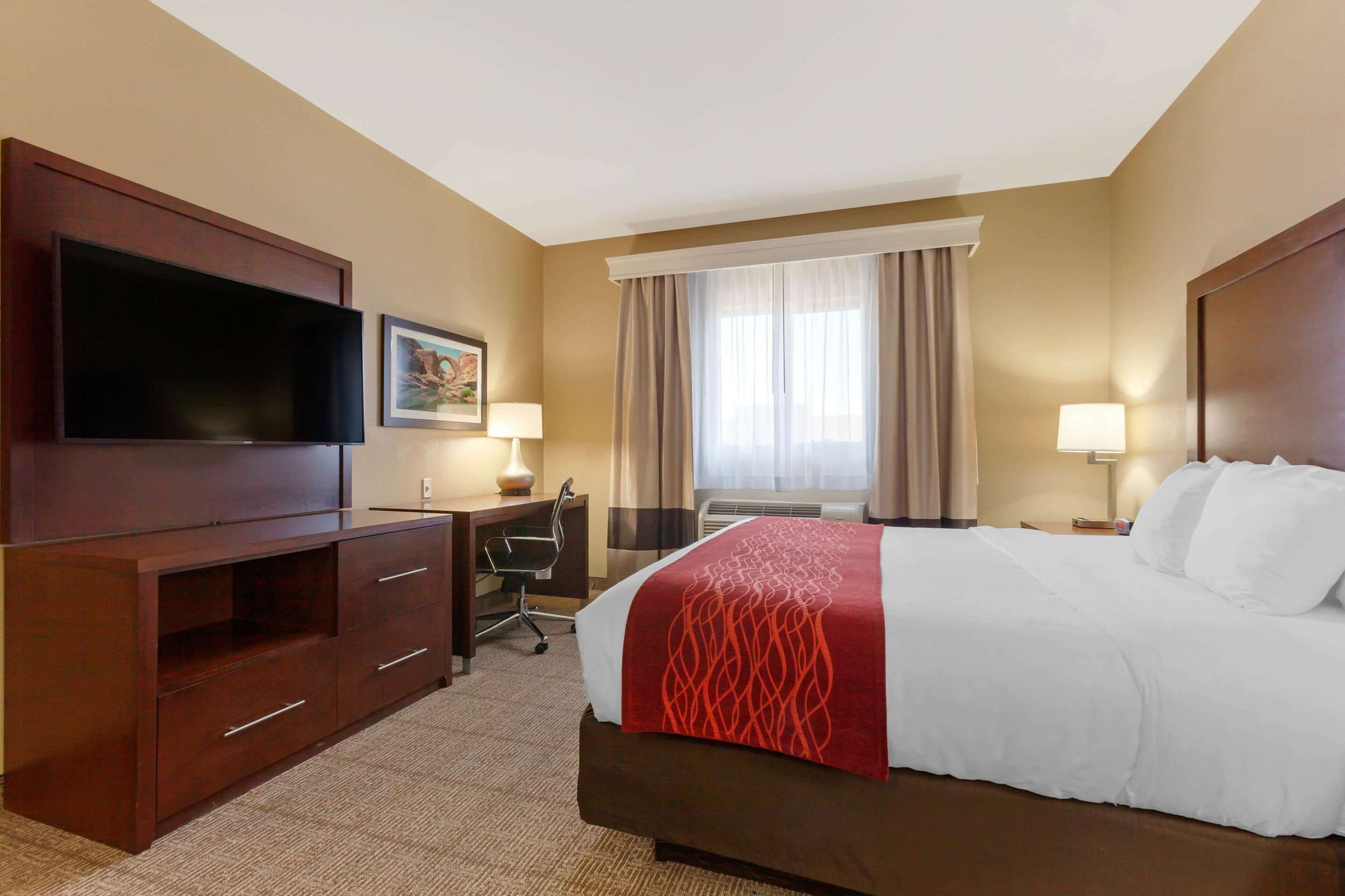 A hotel room with a large bed, a desk with a chair, a flat-screen TV, and a window with curtains.