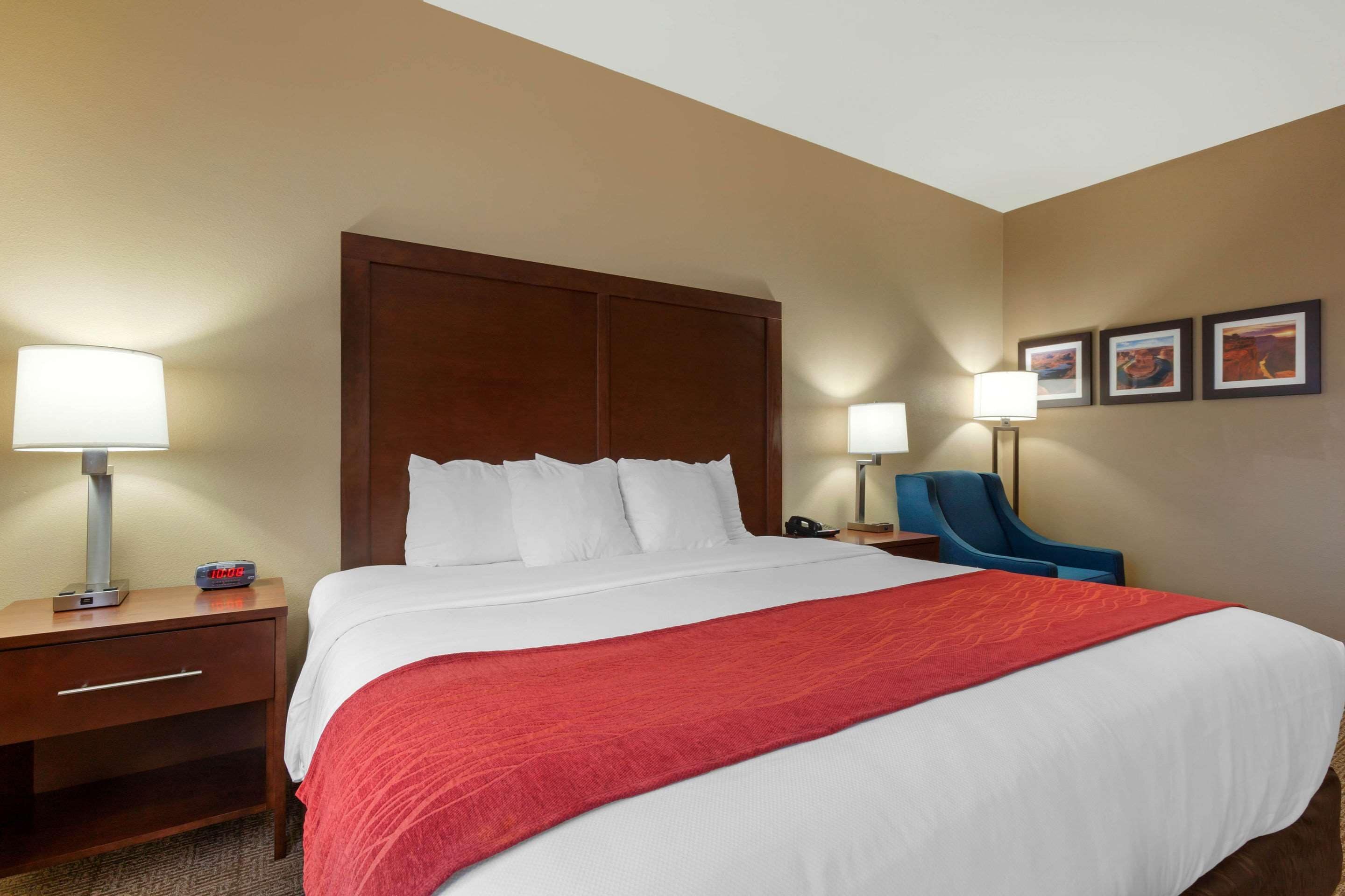 A hotel room with a large bed, a wooden headboard, a red throw blanket, a nightstand with a lamp, a chair, and framed pictures on the wall.