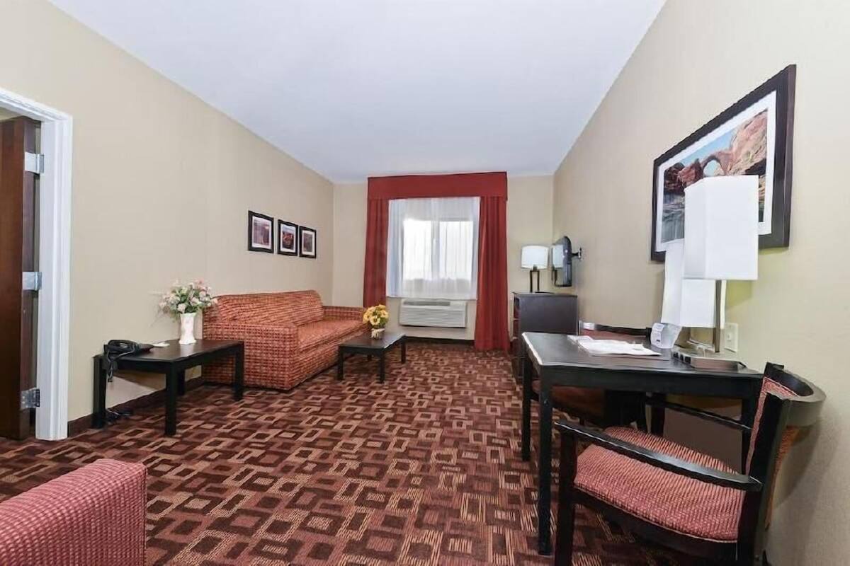 A hotel room with a patterned carpet, a sofa, a small table with a vase, a desk with a chair, a window with red curtains, and framed pictures on the wall.