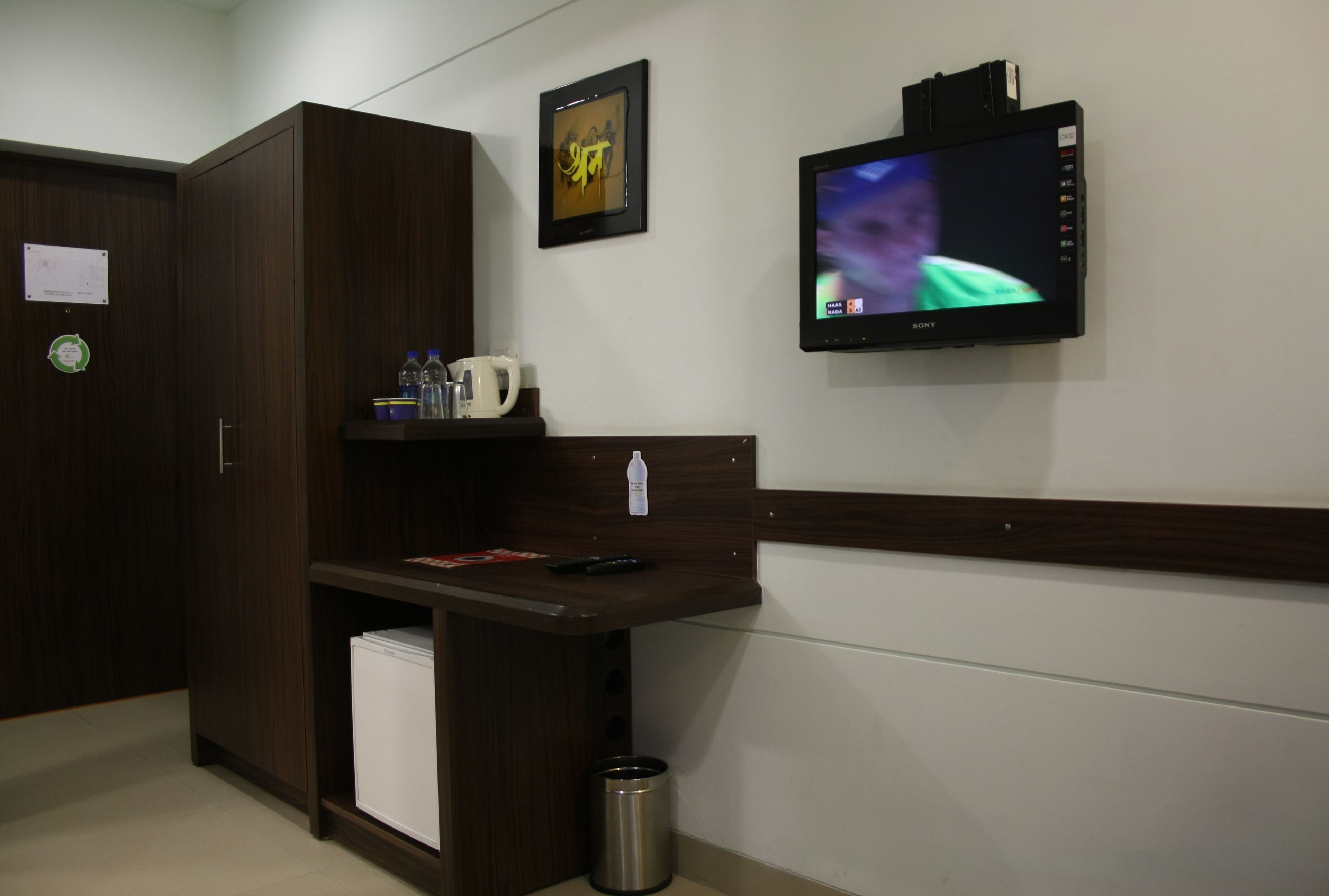 A hotel room with a wooden wardrobe, a small shelf with a coffee maker, a trash bin, and a wall-mounted TV displaying a person.