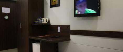 A hotel room with a wooden wardrobe, a small shelf with a coffee maker, a trash bin, and a wall-mounted TV displaying a person.