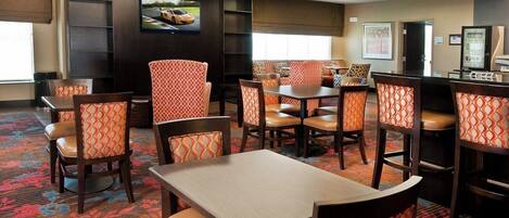 A hotel dining area with multiple tables and chairs, a television mounted on the wall, and a vending machine.