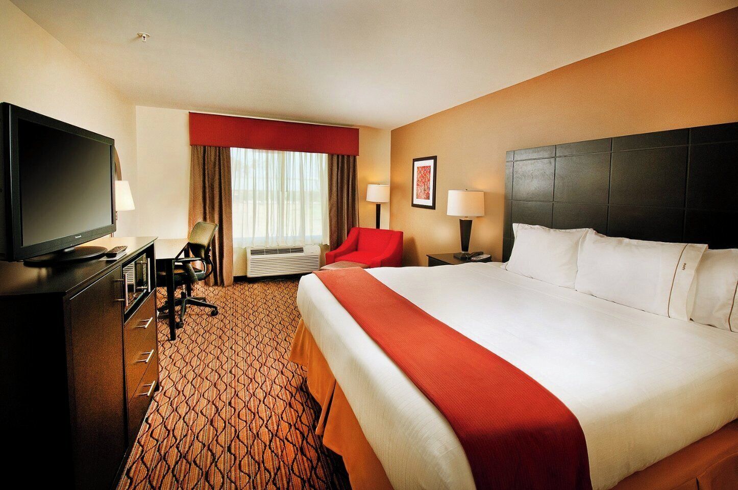A hotel room with a large bed, a desk with a television, a chair, a red accent chair, two bedside lamps, and a framed picture on the wall.