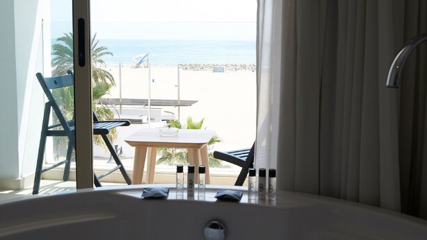 A room with a view of a beach, a table with chairs, and a bathtub with bottles. - Hotel Boutique Sabbia Valencia (Valencia)