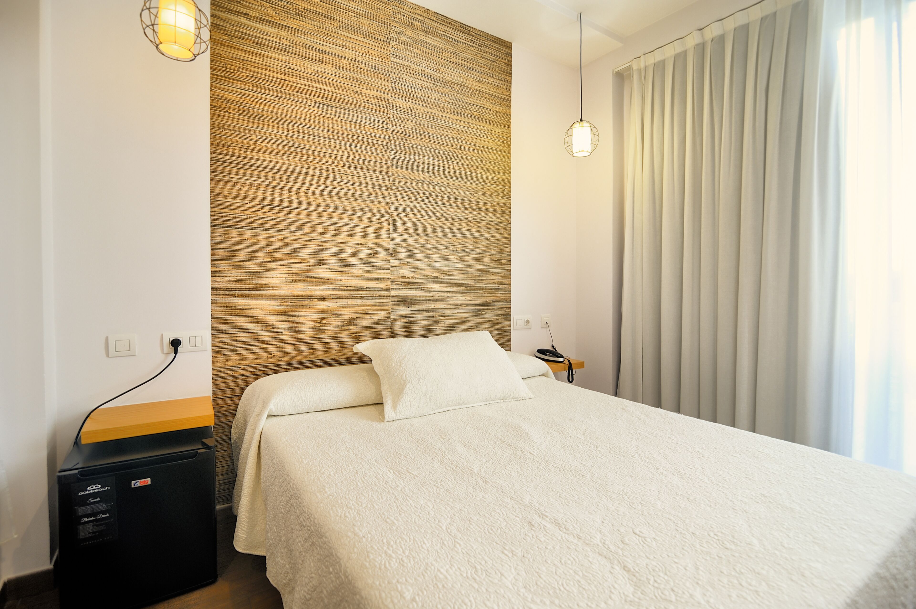 Un lit bien fait, recouvert d’une couvre-lit blanche, avec un mur en bois à texture derrière et une fenêtre avec des rideaux sur le côté droit.