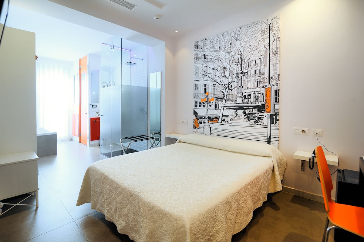 Ein modernes Hotelzimmer mit einem großen Bett, einer Glastür, die auf einen Balkon führt, und einem Stadtbild-Wandgemälde.