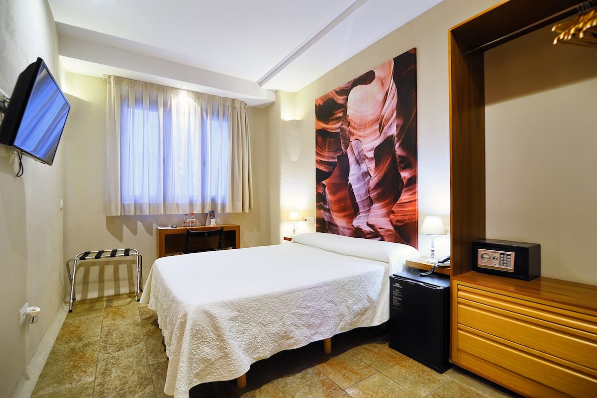 Ein Hotelzimmer mit einem Bett, einem an der Wand befestigten Fernseher, einem Fenster mit Vorhängen, einem großen Gemälde an der Wand und einem Schreibtisch mit Lampe.