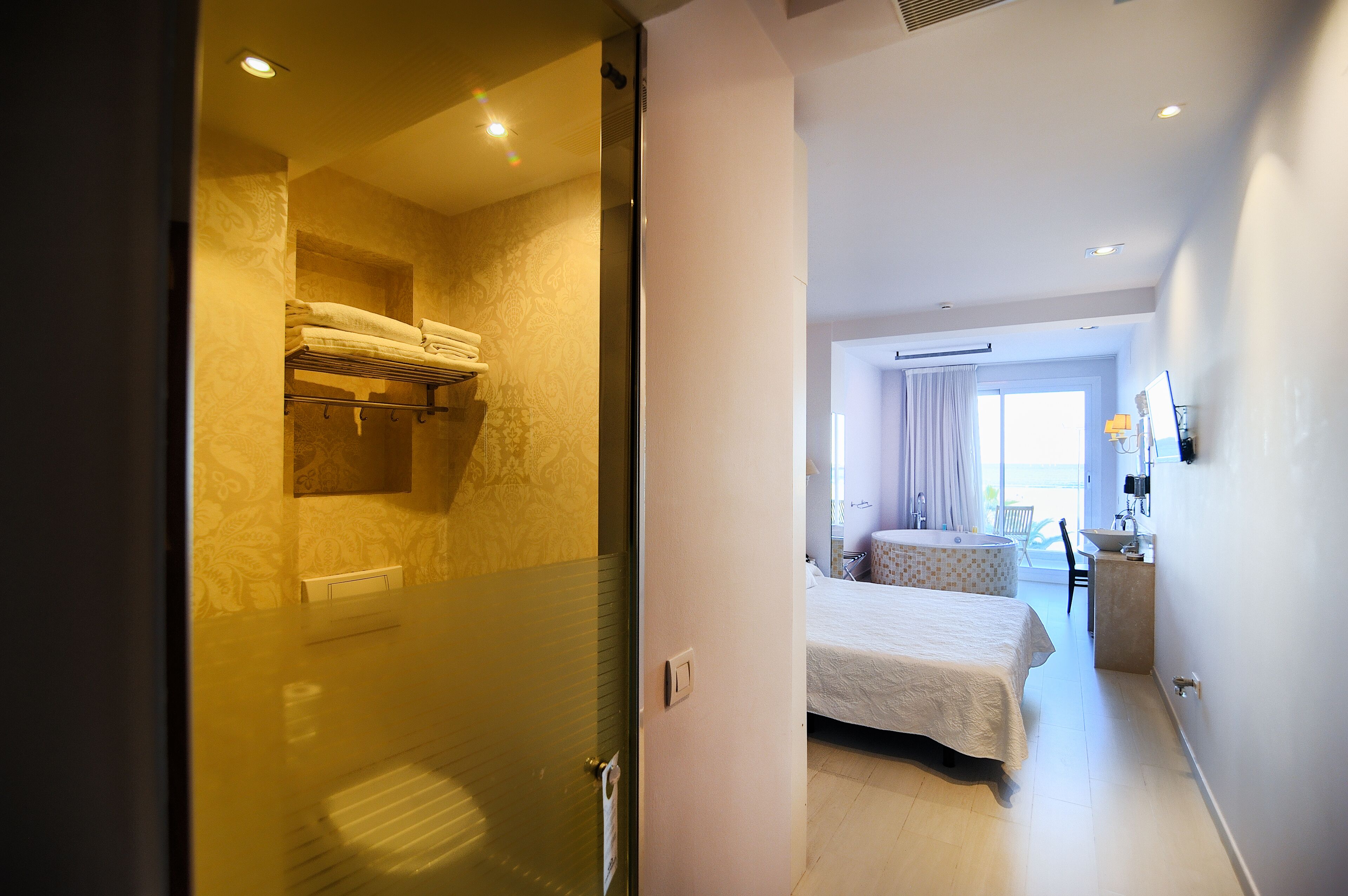 A hotel room with a bed, a small sitting area with a table and chair, and a bathroom with a shower and towel rack.