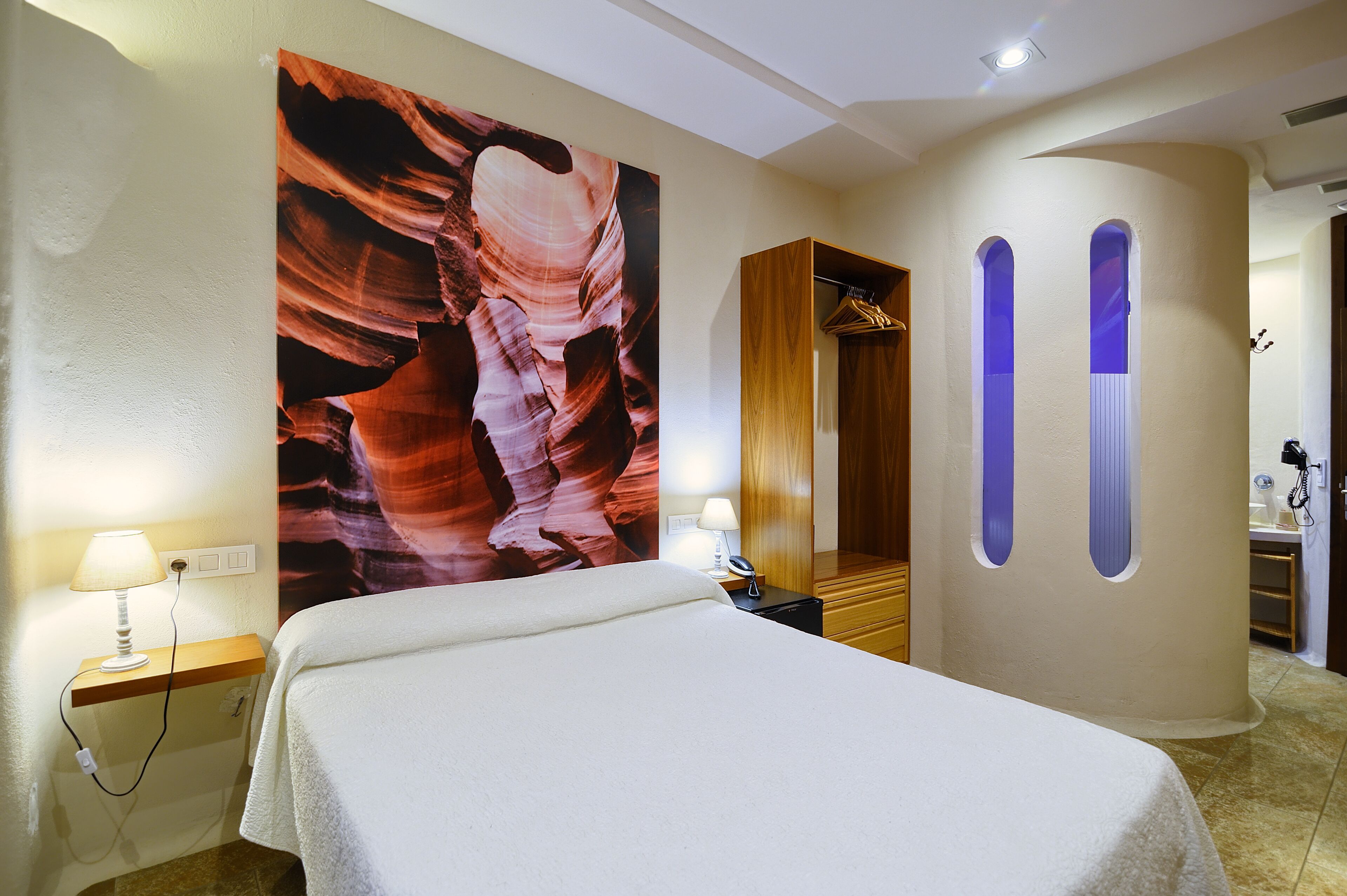 A hotel room with a bed, a large wall painting, a wooden wardrobe, and a bathroom visible through an arched doorway.