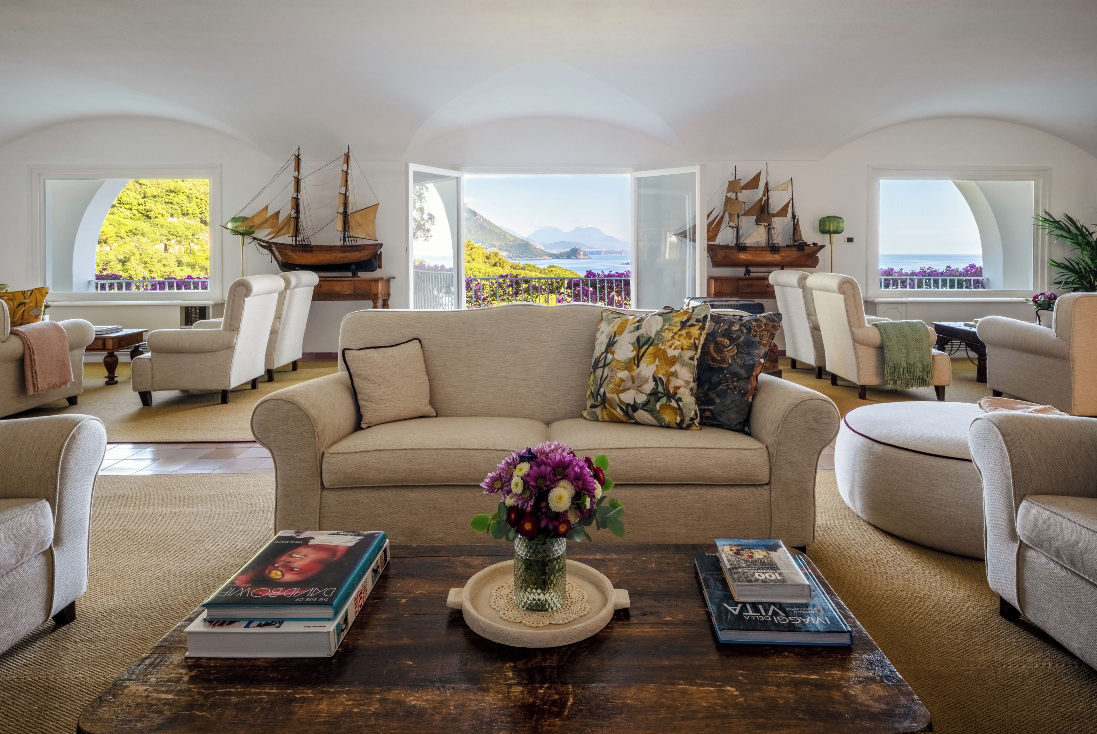 A spacious living room with a large sofa, armchairs, and a coffee table with books and a vase of flowers.