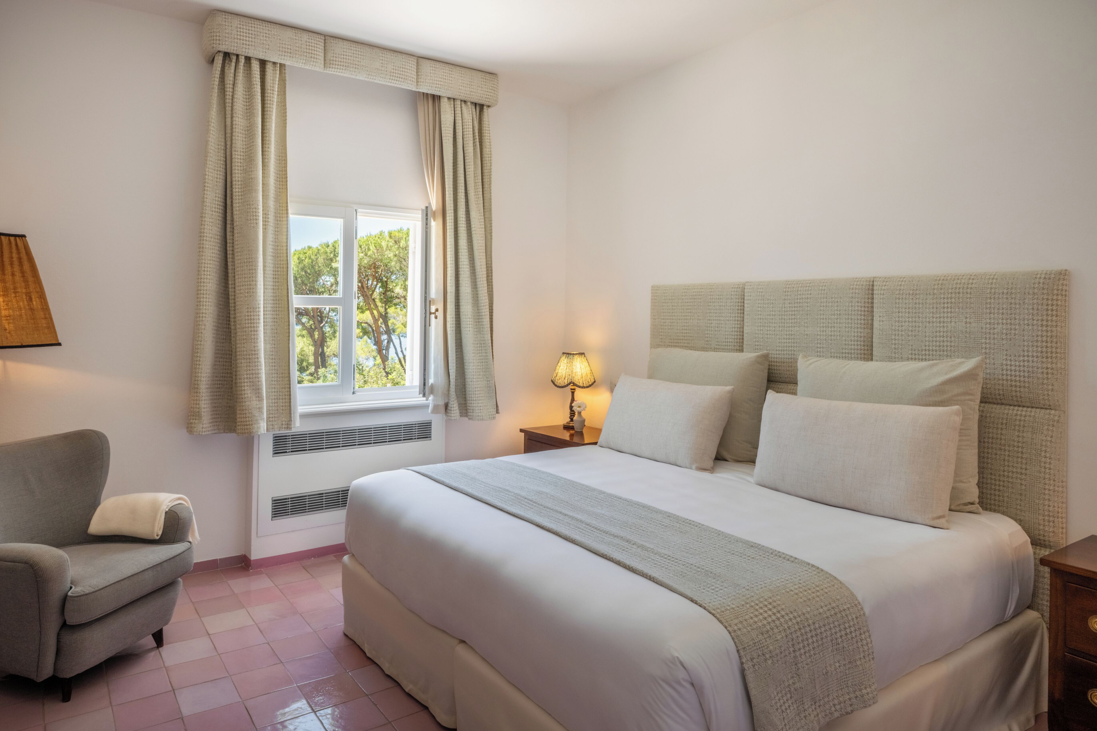 A bedroom with a bed, a chair, a nightstand with a lamp, and a window with curtains.