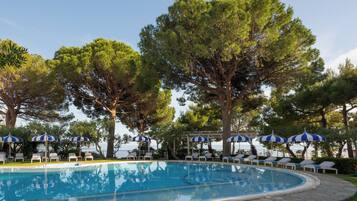 A swimming pool with lounge chairs and umbrellas, surrounded by large trees.