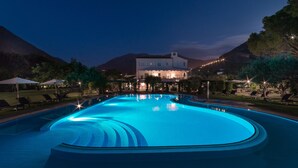 A swimming pool with a curved edge, surrounded by lounge chairs and umbrellas, with a building in the background. - Il Santavenere (Maratea)