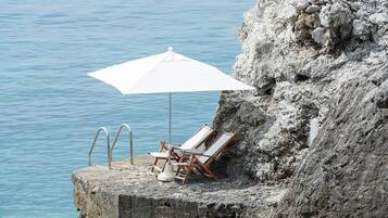 A concrete platform with a white umbrella and two lounge chairs by a rocky cliff overlooking calm water.