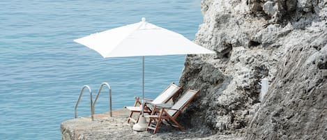 Une plateforme en béton, surmontée d’un parasol blanc et agrémentée de deux chaises longues, est installée au bord d’une falaise rocheuse surplombant une étendue d’eau calme.