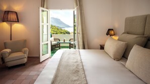 A hotel room with a large bed, a chair, a table with chairs, a lamp, and a view of a mountain and water through an open door. - Il Santavenere (Maratea)