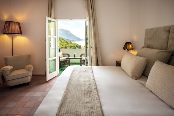 A hotel room with a large bed, a chair, a table with chairs, a lamp, and a view of a mountain and water through an open door. - Il Santavenere (Maratea)