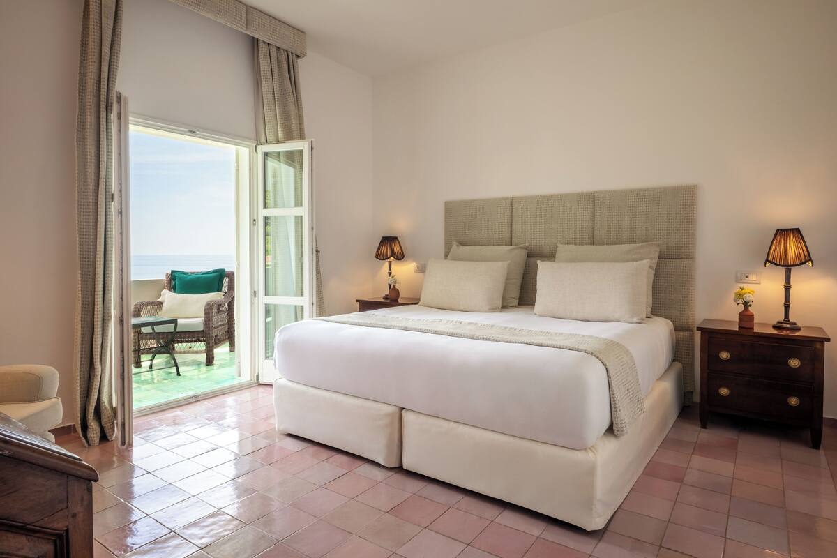 A bedroom with a large bed, two bedside tables, a chair, and a view of the outdoors.