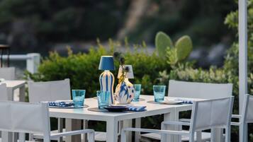 Un'area pranzo all'aperto con tavoli e sedie bianche, bicchieri di vetro blu e un vaso bianco e blu.